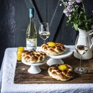 Drei geflochtene Brötchen mit Eigelb auf weißen Ständern, begleitet von einem Glas Weißwein und einer Vase mit frischen Blumen.