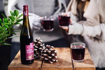 Eine Flasche Roten Glühwein steht auf einem Holztisch, umgeben von Tannenzapfen und dampfenden Gläsern.