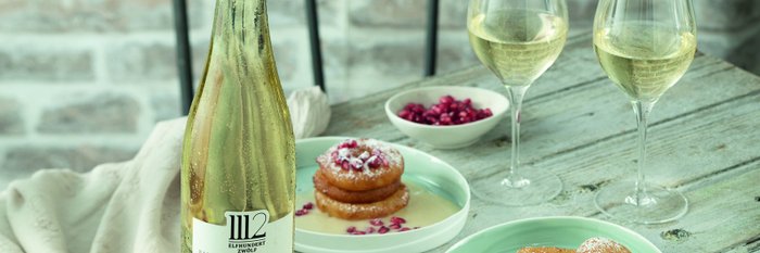 Eine Flasche Grauburgunder steht neben zwei Gläsern Wein und köstlichen Apfelküchle auf einem rustikalen Holztisch.