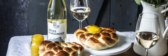Drei geflochtene Brötchen mit Eigelb auf weißen Ständern, begleitet von einem Glas Weißwein und einer Vase mit frischen Blumen.