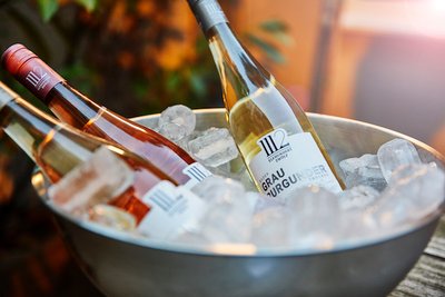 Drei Flaschen Wein, darunter ein Grau Burgunder, liegen in einem silbernen Eiskübel mit vielen Eiswürfeln.