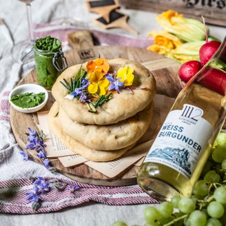 Zwei Gläser Weißwein stehen neben köstlichen Fladenbroten, dekoriert mit bunten Blüten und frischen Kräutern auf einem Tisch.