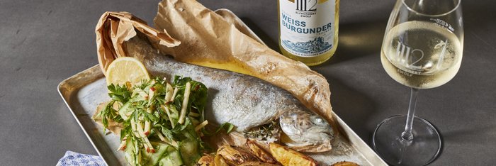 Ein Teller mit gebratenem Fisch, knusprigen Kartoffeln und einem frischen Salat aus Gurken und Kräutern, garniert mit Zitrone.