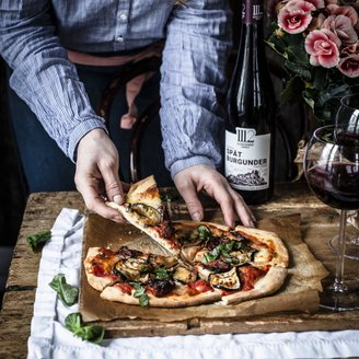 Eine Person greift nach einem Stück Pizza, die mit frischem Gemüse und Kräutern belegt ist, auf einem rustikalen Holztisch.