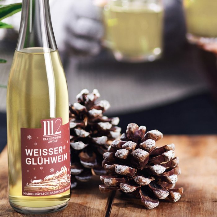 Eine Flasche Weißer Glühwein steht auf einem Holztisch, umgeben von Tannenzapfen und dampfenden Gläsern in den Händen von Personen.