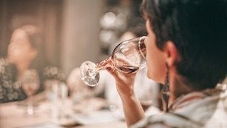 Eine Frau mit kurzem Haar genießt einen Schluck Rotwein aus einem eleganten Glas, während sie in geselliger Runde sitzt.