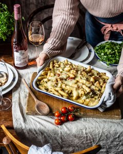 Eine Person hält eine Auflaufform mit Käse überbackenen Nudeln, umgeben von frischen Tomaten und einem Glas Roséwein.