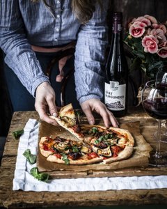 Eine Person greift nach einem Stück Pizza, die mit frischem Gemüse und Kräutern belegt ist, auf einem rustikalen Holztisch.
