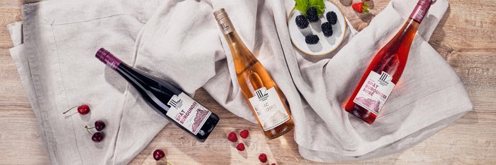 Drei Flaschen Wein liegen auf einem hellen Tuch, umgeben von frischen Beeren und einer kleinen Schale mit schwarzen Johannisbeeren.
