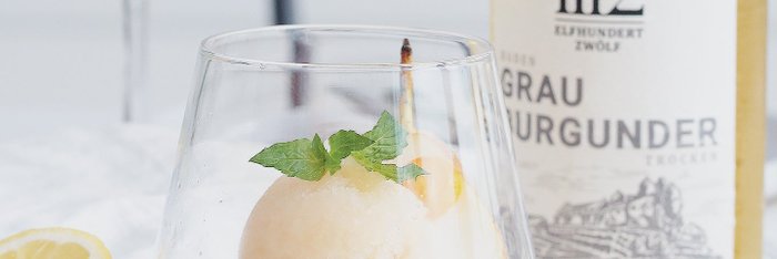 Das Sorbet in einem Kristallglas wird von Zitronen- und Kumquatstücken sowie einem Glas Weißwein begleitet.
