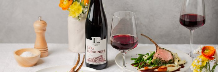 Zwei kunstvoll angerichtete Teller mit Lamm und Gemüse, begleitet von einem Glas Rotwein und einer stilvollen Blumendekoration.