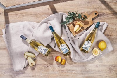 Drei Flaschen Weißwein liegen auf einem hellen Tuch, umgeben von frischen Äpfeln und einer Zitrone auf einem Holzbrett.