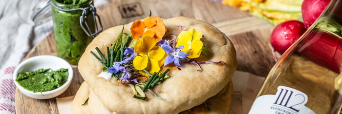 Zwei Gläser Weißwein stehen neben köstlichen Fladenbroten, dekoriert mit bunten Blüten und frischen Kräutern auf einem Tisch.