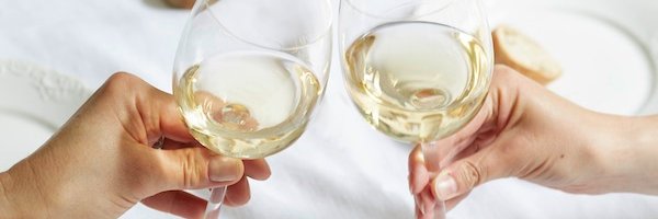 Zwei Hände stoßen mit Gläsern voll Weißwein an, umgeben von einem eleganten Tischsetting mit Besteck und Geschirr.