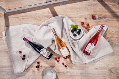 Drei Flaschen Wein liegen auf einem hellen Tuch, umgeben von frischen Beeren und einer kleinen Schale mit schwarzen Johannisbeeren.