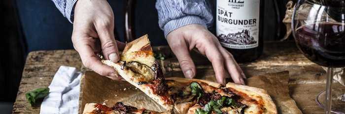 Eine Person greift nach einem Stück Pizza, die mit frischem Gemüse und Kräutern belegt ist, auf einem rustikalen Holztisch.