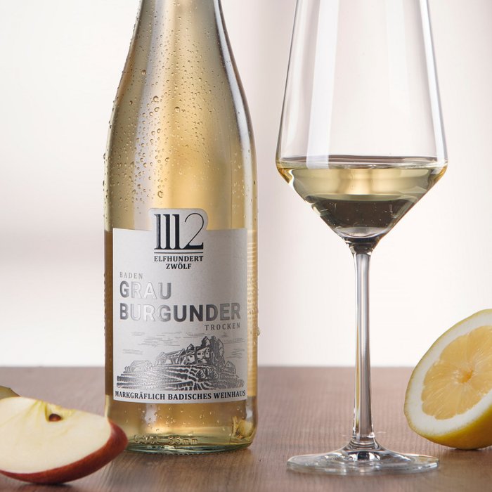 Eine Flasche 1112 Grauburgunder steht neben einem mit Wein gefüllten Glas, während frische Äpfel und eine Zitrone daneben auf einem Holztisch liegen.