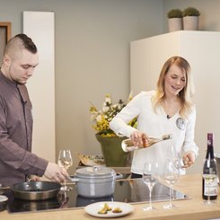 Ein Mann und eine Frau bereiten in einer modernen Küche ein Essen vor, während die Frau Wein in Gläser einschenkt.
