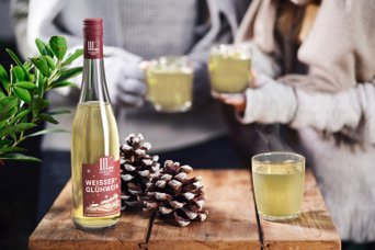 Auf einem rustikalen Tisch liegen Tannenzapfen, neben einer Flasche Glühwein und einem Glas mit warmem Getränk.