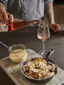 Auf dem Tisch steht ein frisch gebackenes Gericht mit Äpfeln und Rosinen, daneben eine Schüssel mit Vanillesoße und ein Glas Roséwein wird eingeschenkt.