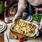 Eine Person hält eine Auflaufform mit Käse überbackenen Nudeln, umgeben von frischen Tomaten und einem Glas Roséwein.