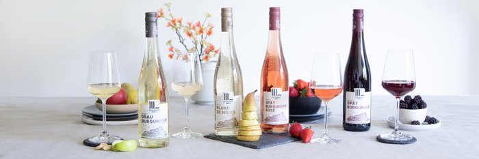 Auf einem Tisch stehen vier Weinflaschen, umgeben von frischen Früchten und eleganten Gläsern, die eine festliche Atmosphäre schaffen.