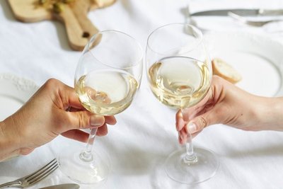 Zwei Hände stoßen mit Gläsern voll Weißwein an, umgeben von einem eleganten Tischsetting mit Besteck und Geschirr.
