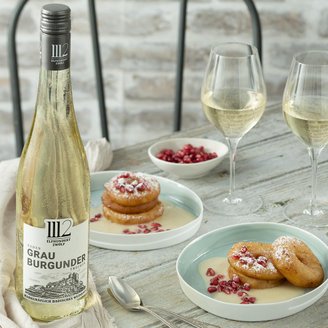 Eine Flasche Grau Burgunder steht neben zwei Gläsern Wein und zwei Tellern mit süßen, mit Puderzucker bestäubten Apfelküchlein.