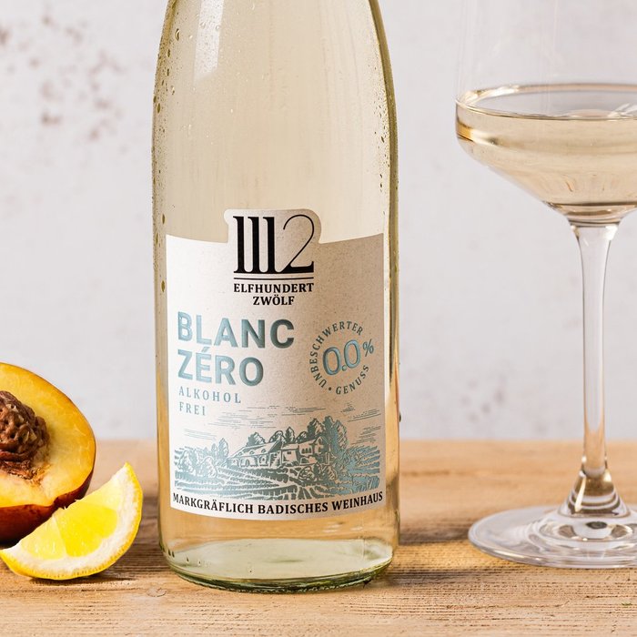 Auf einem rustikalen Tisch sind eine Flasche Blanc Zéro und ein Glas mit hellem Wein, umgeben von frischen Früchten, platziert.