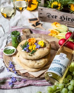 Zwei Gläser Weißwein stehen neben köstlichen Fladenbroten, dekoriert mit bunten Blüten und frischen Kräutern auf einem Tisch.