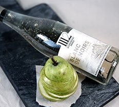 Eine Flasche Blanc de Noirs Wein liegt auf einem schwarzen Brett, neben einer kunstvoll geschnittenen grünen Birne.