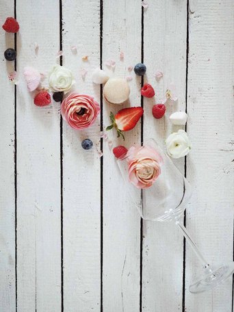 Ein elegantes Glas liegt auf einem weißen Holztisch, umgeben von frischen Blumen und bunten Beeren, die eine festliche Stimmung erzeugen.