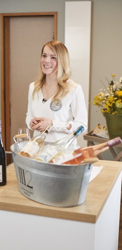 Eine lächelnde Frau in einem weißen Oberteil steht an einem Tisch mit einer Schalle gefüllt mit Eis, Weinflaschen und Gläsern.