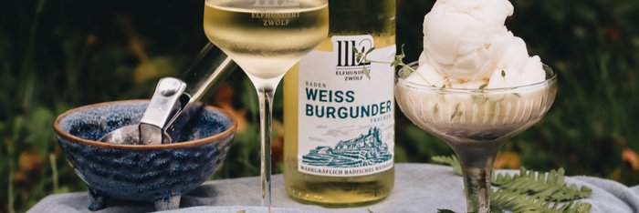 Ein Glas mit Weißwein und eine Flasche stehen neben einer eleganten Schale mit Sorbet, alles auf einem blauen Tuch drapiert.