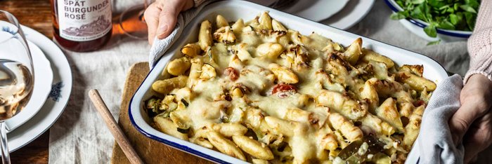 Eine Person hält eine Auflaufform mit Käse überbackenen Nudeln, umgeben von frischen Tomaten und einem Glas Roséwein.