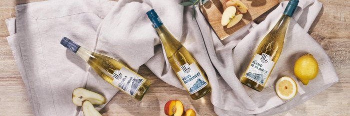Drei Flaschen Weißwein liegen auf einem hellen Tuch, umgeben von frischen Äpfeln und einer Zitrone auf einem Holzbrett.