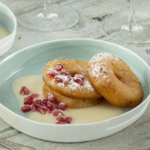 Zwei goldbraune, frittierte Teigringe liegen auf einer hellblauen Schale, garniert mit Puderzucker und Granatapfelkernen.