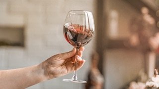 Eine Hand präsentiert ein Weinglas mit rotem Wein, das Licht reflektiert sich auf der Glasoberfläche und schafft eine festliche Stimmung.