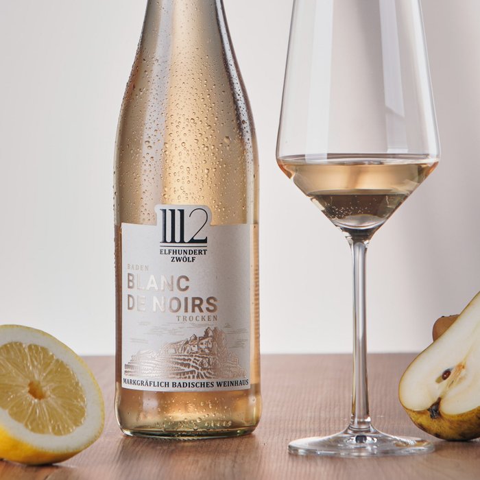 Eine Flasche 1112 Blanc de Noirs steht neben einem mit Wein gefüllten Glas, während eine frische Birne und eine Zitrone daneben auf einem Holztisch liegen.