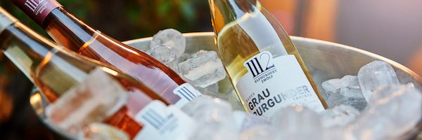 Drei Flaschen Wein, darunter ein Grau Burgunder, liegen in einem silbernen Eiskübel mit vielen Eiswürfeln.