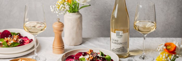 Ein eleganter Tisch gedeckt mit einem bunten Salat, einem Glas Weißwein und einer Flasche Grau Burgunder, umgeben von Blumen.