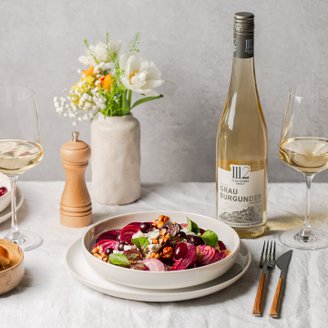 Ein eleganter Tisch gedeckt mit einem bunten Salat, einem Glas Weißwein und einer Flasche Grau Burgunder, umgeben von Blumen.