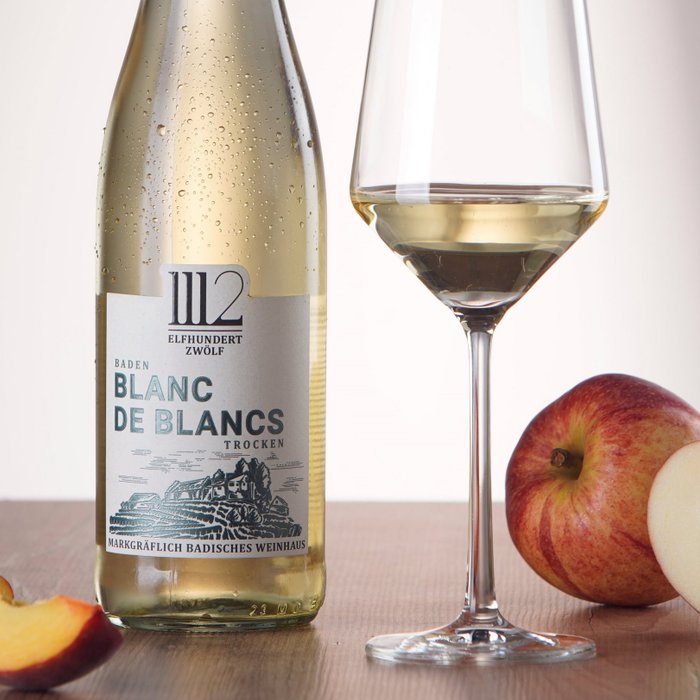 Eine Flasche 1112 Blanc de Blancs steht neben einem mit Wein gefüllten Glas, während frische Äpfel und Pfirsiche daneben auf einem Holztisch liegen.