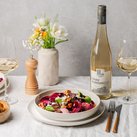 Ein eleganter Tisch gedeckt mit einem bunten Salat, einem Glas Weißwein und einer Flasche Grau Burgunder, umgeben von Blumen.