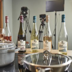 Fünf Flaschen Wein stehen auf einem Tisch, umgeben von Weingläsern und Kochgeschirr in einer modernen Küche.