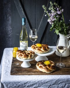 Drei geflochtene Brötchen mit Eigelb auf weißen Ständern, begleitet von einem Glas Weißwein und einer Vase mit frischen Blumen.