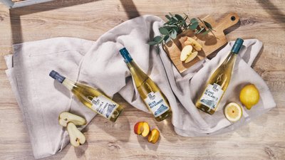 Drei Flaschen Weißwein liegen auf einem hellen Tuch, umgeben von frischen Äpfeln und einer Zitrone auf einem Holzbrett.