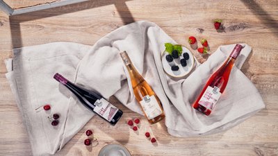 Drei Flaschen Wein liegen auf einem hellen Tuch, umgeben von frischen Beeren und einer kleinen Schale mit schwarzen Johannisbeeren.