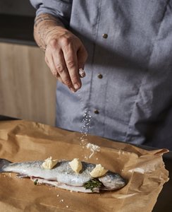 Eine Hand streut Salz über einen frischen Fisch, der auf Backpapier mit Butter und Kräutern vorbereitet wird.