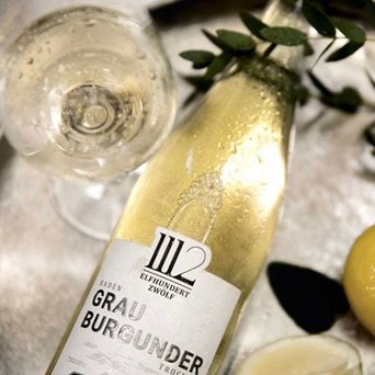 Auf einem Tisch liegen eine Flasche Grau Burgunder und ein Glas, umgeben von Zitronenscheiben und dekorativen Blättern.
